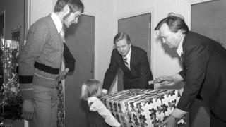 Tyttö saa vierailta ison lahjapaketin. Väki on juhlavaatteissa.