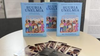 vaalealla pyöreällä pöydällä esillä Suuria unelmia kirjoja. Kirjan kannessa sinisellä taustalla kirjan otsikko ja  animoituja maahanmuuttotaustaisia henkilöitä