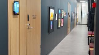 Käytävä. Vasemmalla tumma seinä, jossa vaaleita ovia. Ovien välissä seinällä piirrustuksia.