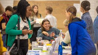 Kuvassa osallistujat keskustelevat ständipöydän äärellä vuoden 2025 Tervetuloa Vantaalle -tapahtumassa Koivukylässä. Taustalla nuoret pelaavat UNO-korttipeliä rennossa tunnelmassa.