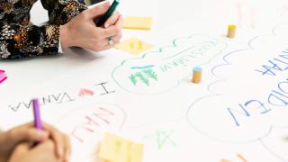 Kuvassa ideoidaan yhdessä pöydän ympärillä. Kuvassa näkyy ihmisten käsiä sekä yhdessä värikkäillä tusseilla kirjoitettuja ideoita ja tekstejä, kuten I (sydän) Vantaa. värikkäitä 