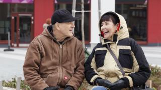 vanhus pariskunta Paksuihin takkeihin pukeutuneet mies ja nainen istuvat penkillä vierekkäin toisilleen hymyillen.