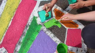 Asvalttiin maalattuja värikkäitä ja kulmikkaita muotoja. Kuvan vasemmassa laidassa näkyy kahden ihmisen kädet, jotka maalaavat muotoja vihreään ja turkoosiin väriin kastetuilla pensseleillä.