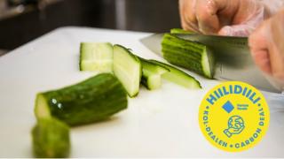 Kurkkuja leikataan valkoisella leikkauslaudalla. Hiilidiili logo.