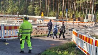 Rakentaja katsoo kun vanhemmat ja lapsi kävelevät kohti suojatietä työmaa-alueen vieressä.