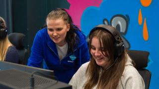 Nuori pelaa tietokoneella, ohjaaja katsoo vieressä. 