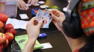 Henkilö pelaa korttipeliä. Taustalla näkyy muiden pelaajien käsiä.