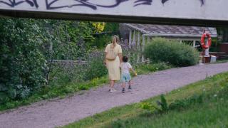 Kesäisiin vaatteisiin pukeutunut aikuinen ja taapero kävelevät soratiellä.