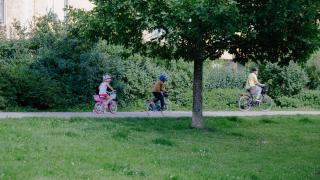 Kaksi lasta pyöräilee aikuisen perässä puistossa Aviapoliksen suuralueella.