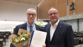 Vantaan Voimisteluseura ry:n jäsen Seppo Nieminen ja kaupunginjohtja Pekka Timonen seisovat vierekkäin valtuustosalissa. Niemisellä käsissään kunniakirja ja kukkakimppu.