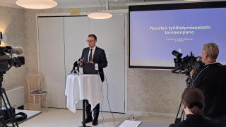 Työministeri Marttinen seisoo mediatilaisuudessa huoneen etuosassa ja kertoo medialle uudesta tukimallista.