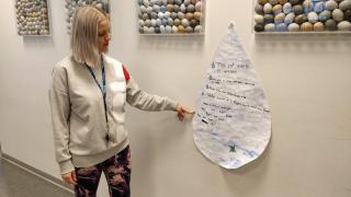 Ida seisoo koulun käytävällä ja osoittaa seinällä olevaa vesipisaranmuotoista julistetta.