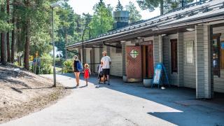 Ulkoilumaja ulkoa päin kuvattuna. Pääovi on auki ja sen edessä kävelee lapsiperhe kesävaatteissa.