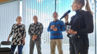 Kuvassa on neljä henkilöä, joista etualalla oleva puhuu mikrofoniin.