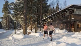 Kaksi miestä uimashortseissa, pipoissa ja uimatossuissa kävelemässä Kuusijärven sähkösaunalta kohti avantoa lumisessa maisemassa.