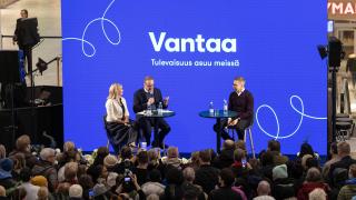 Presidentin haastattelu menossa lavalla sisätiloissa. Edessä on yleisöjoukko katsomassa ja takana seinäke, jossa lukee Vantaa..