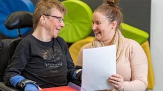 Pyörätuolissa istuva mies ja nainen hymyilevät