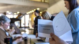 Äänestyslippu vaalisimulaatiossa 30.10.2024 ja taustalla äänestäviä ihmisiä.