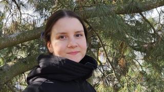 Mustatakkinen nainen seisoo havupuun edessä.