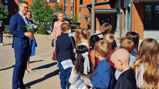Presidentti Stubb seisoo kukat kädessään päiväkodin pihalla. Lapset heiluttavat Suomen lippuja.
