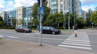 Lapsi ylittää suojatien potkulaudalla, kaksi auto odottaa liikennevaloissa. Taustalla vaaleita kerrostaloja.