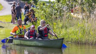 Kaksi aikuista ja yksi lapsi melomassa kesäisessä rantamaisemassa.