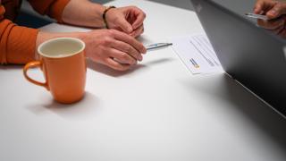 Kuvaan rajautuu ihmisen kädet, toisen ihmisen kädet tietokoneella. Kuva havainnollistaa asiointitilannetta.