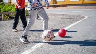 Kahden lapsen jalat, jalkapallo ja punainen pallo pihamaalla.