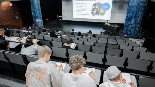 Opiskelijat istuvat auditoriossa tekemässä muistiinpanoja. Valkokankaalla näkyy Hybridikeskuksen esittelyteksti.
