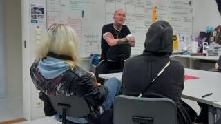 Tatuoija Olli Ruikka seisoo huoneessa puhumassa nuorille, jotka istuvat tuoleilla selkä kameraan päin.