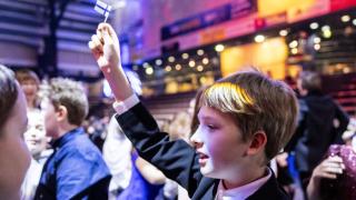 Pojalla on puku päällään ja pieni Suomen lippu kohotettuna kädessään. Taustalla on muita oppilaita Energia Areenan tiloissa.