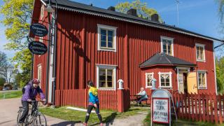 kahvila punaisessa puisessa talossa ja aurinkoisesta päivästä nauttivat ihmiset.