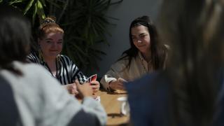 Oppilaitosnuorisotyöntekijät Tiina Selenius ja Milja Heinonen istuvat pöydän ääressä pelaamassa Uno-korttipeliä kahden nuoren kanssa, joista näkyy selkäpuolet.