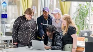 Yksi nuori istuu pöydän ääressä käyttämässä kannettavaa tietokonetta. Hänen ympärillään neljä muuta, hymyilevää nuorta katsomassa tietokoneen näyttöä. Kuvan vasemmassa yläreunassa Vantaan kaupungin, Vipuvoimaa EU:lta - ja EU:n logot.