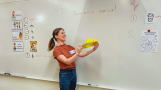 Ensimmäisen luokan opettaja seisoo luokkahuoneen taulun edessä hymyillen. Hän pitää käsissään keltaista lippalakkia. 