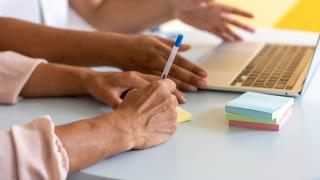 Kädet kirjoittamassa muistilappua ja kannettava tietokone.