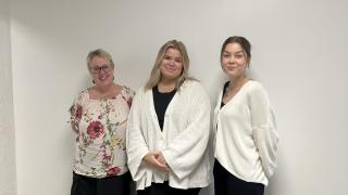 Kolme kouluvalmentajaa seisoo valkoisen seinän edessä. He hymyilevät.