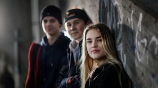 Kolme nuorta seisoo betoniseinään nojaillen ja he katsovat kameraan. 