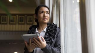 Henkilöllä on tabletti kädessä ja hän katsoo ikkunasta ulos.