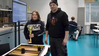 Kuvassa etualalla suuri backgammon-lauta pöydällä, jonka takana Milko kultapokaali käsissään ja vieressä seisoo Don Rahikainen.