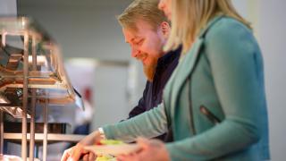 Kuvassa kaksi henkilöä annostelee lautaselle ruokaa linjastosta.