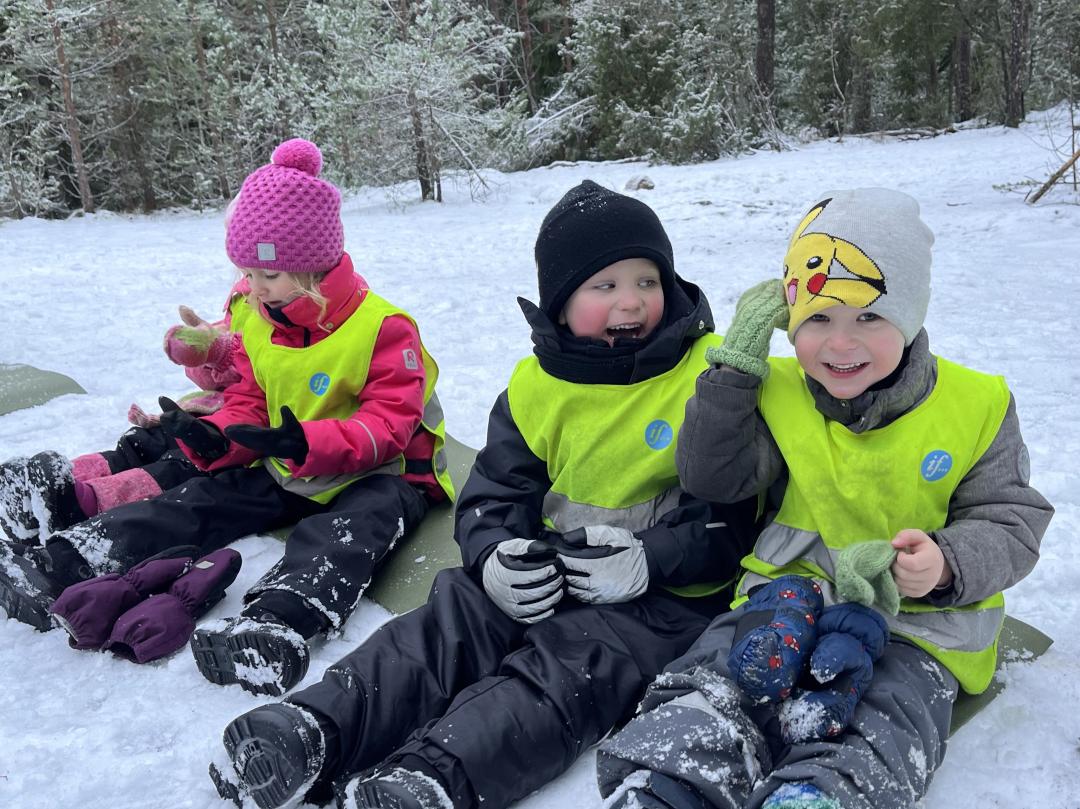 Barnen sitter på den snöiga marken på ett liggunderlag och ler. 