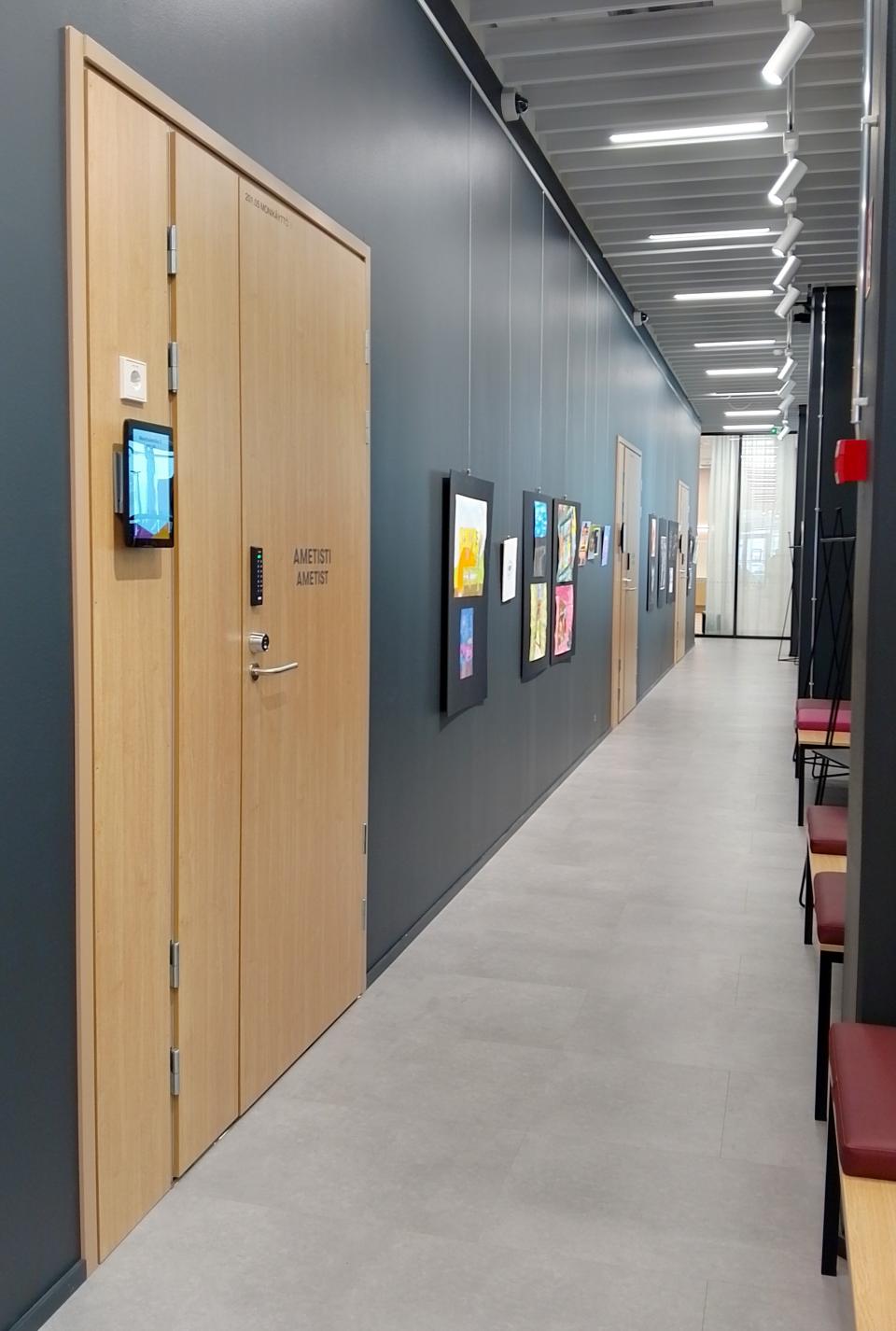 Käytävä. Vasemmalla tumma seinä, jossa vaaleita ovia. Ovien välissä seinällä piirrustuksia.