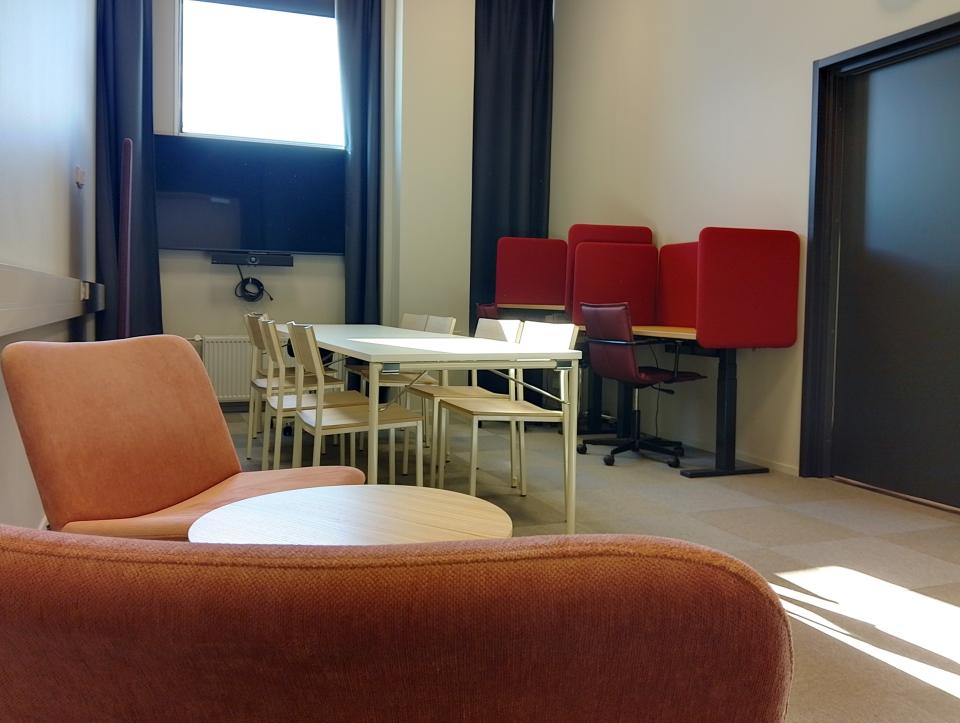 Kapeassa vaaleassa huoneessa pitkäpöytä, tuolit, kaksi työpistettä ja kaksi nojatuolia.Takaseinällä tv ja ikkuna.