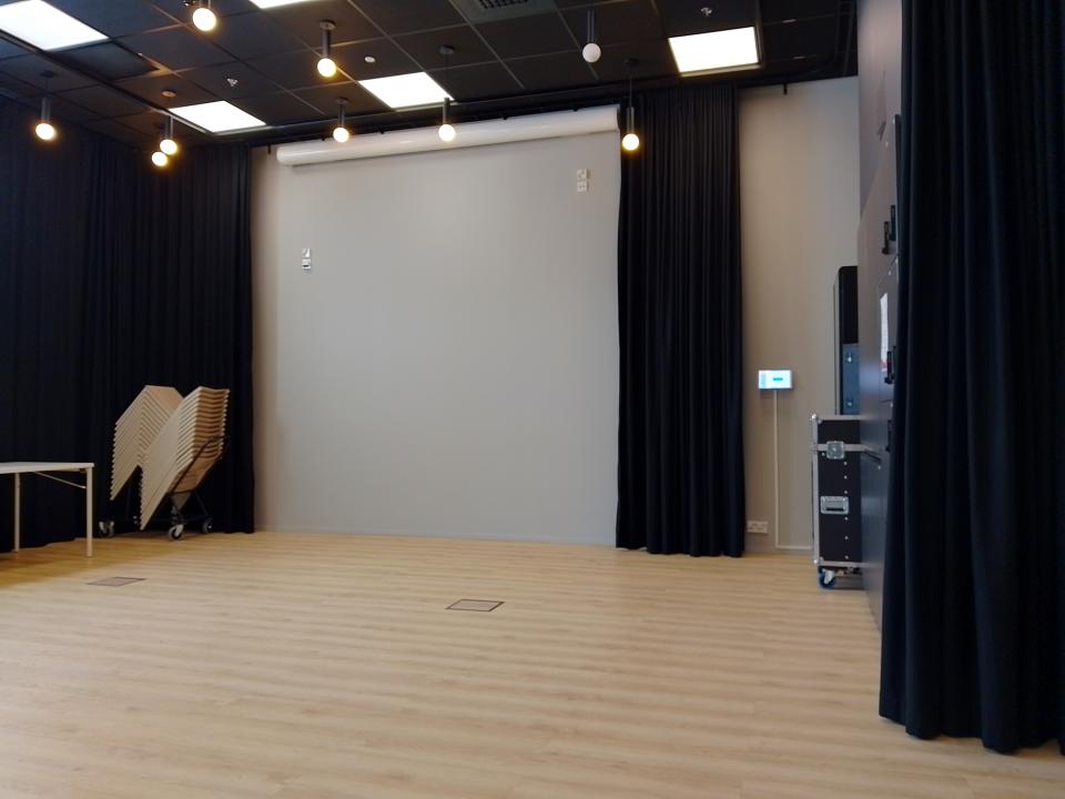 Tyhjätila, jossa vaalea lattia, seiniä kiertää musta verho. Katossa lamppuja ja videotykki.