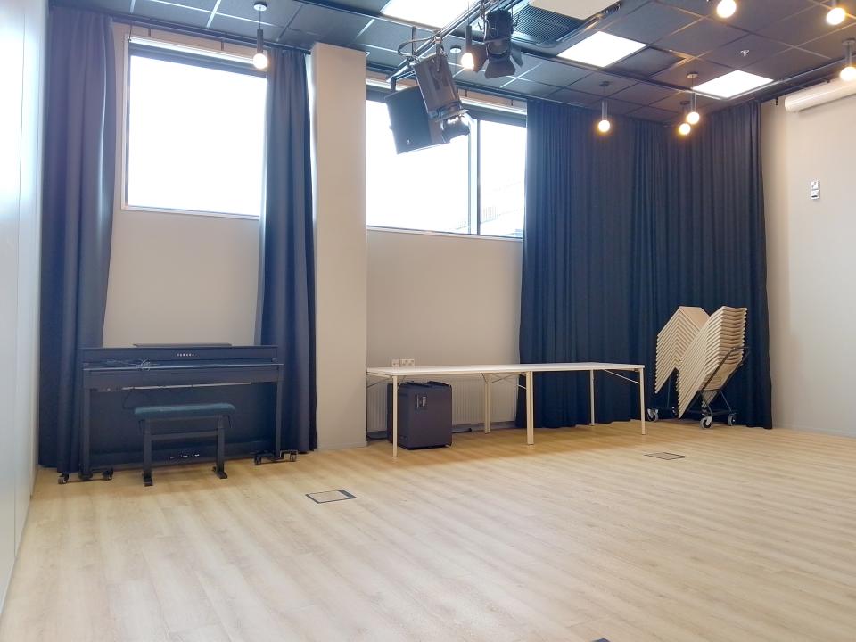 Tyhjätila, jossa vaalea lattia, seiniä kiertää musta verho. Katossa lamppuja. Seinänvieressä pöytiä ja tuoleja pinossa. 