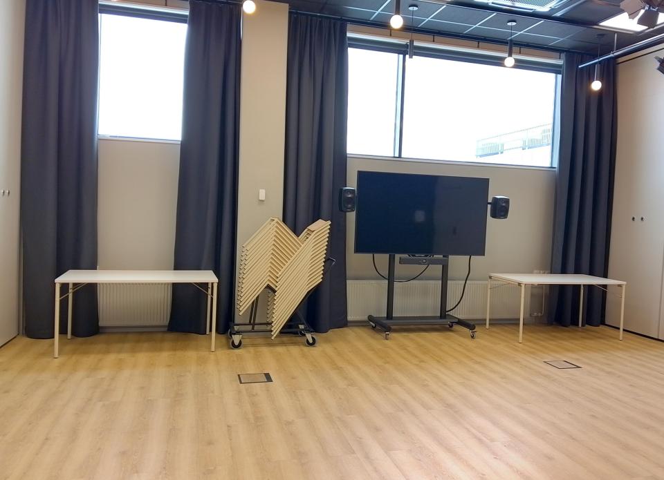 Tilassa tyhjää lattiaa ja seinän vieressä kaksi pöytää, tuoleja ja siirrettävä näyttö.