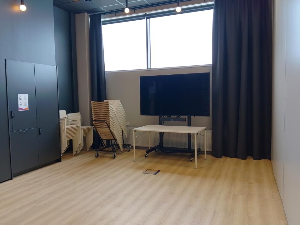 Tyhjätila, jossa vaalea lattia, seiniä kiertää musta verho. Seinän vieressä pinossa tuolleja, pöytä ja iso näyttö. 