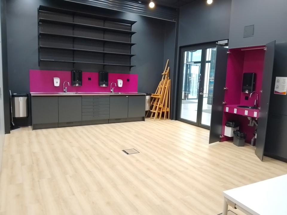 Tyhjätila, jossa vaalea lattia. Tilan perällä vesipisteitä ja seinällä hyllyjä. Oikealla nurkassa lasiovi.