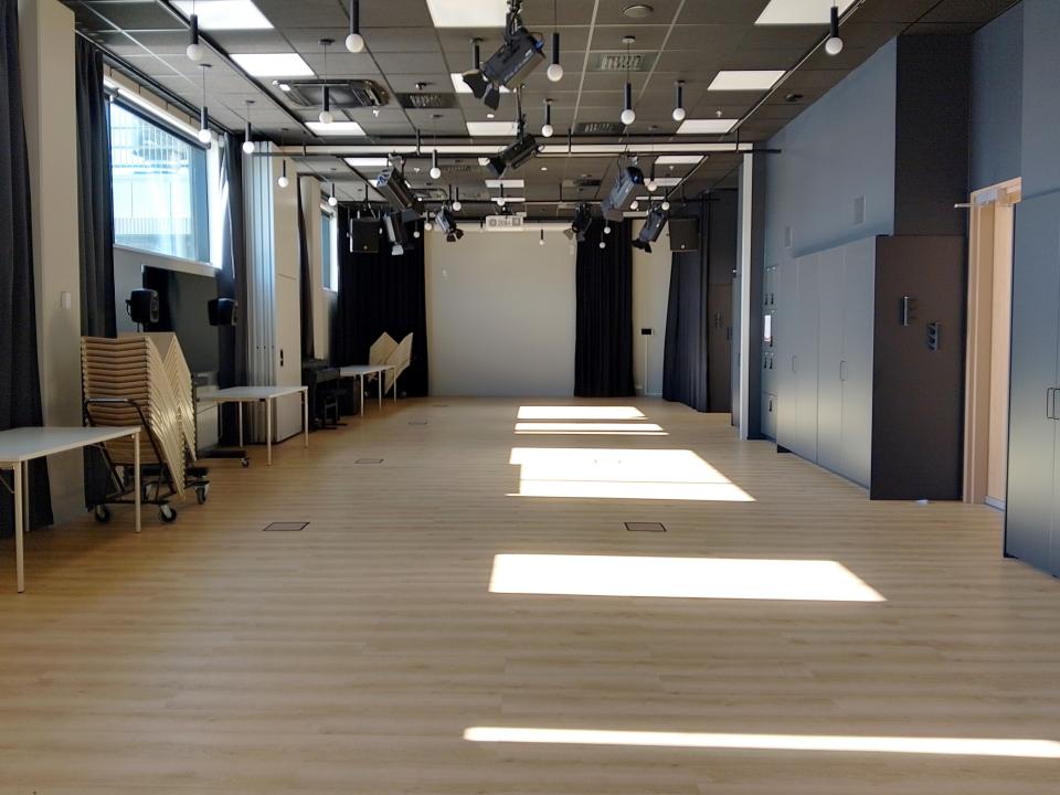 Tyhjätila, jossa vaalea lattia, seiniä kiertää musta verho, seinän vieressä kalusteita pinossa. Katossa lamppuja ja videotykki.